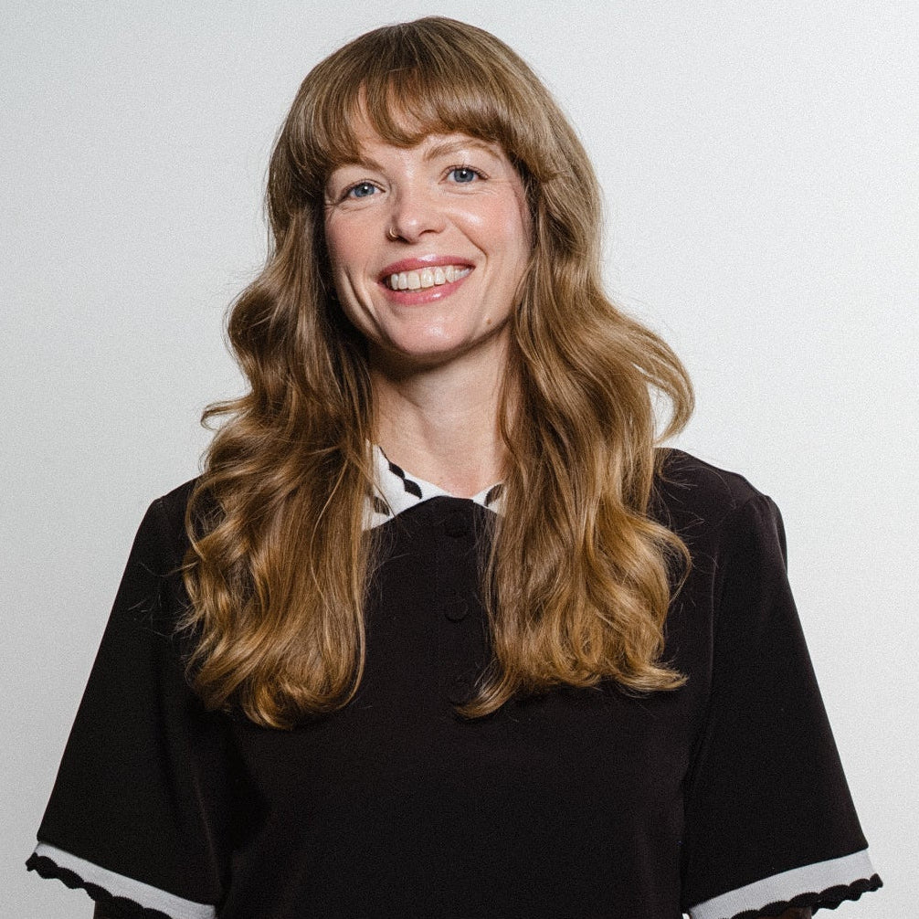 A smiling woman with long wavy light brown hair and visible tattoos on both arms, wearing a black short-sleeve top with white trim at the collar and sleeve cuffs, paired with black pants, standing against a plain light gray wall.