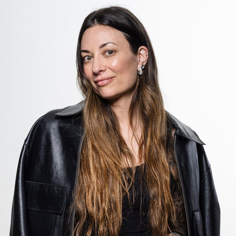 A woman with long brown hair stands wearing a black leather jacket, black top, and black wide-leg pants against a plain light gray wall.